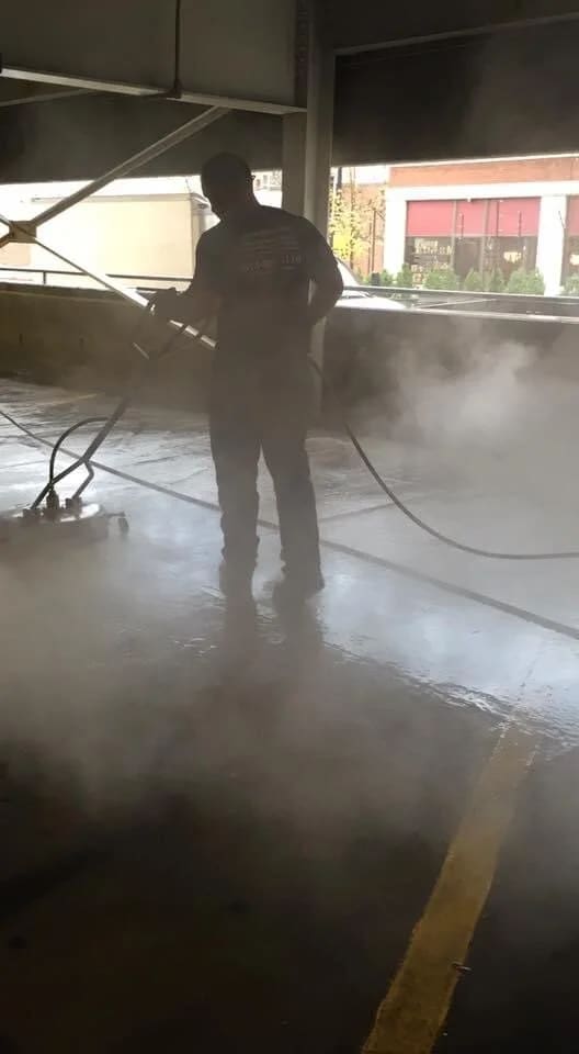 Crew member washing a parking garage floor with hot water, steam rising off the concrete